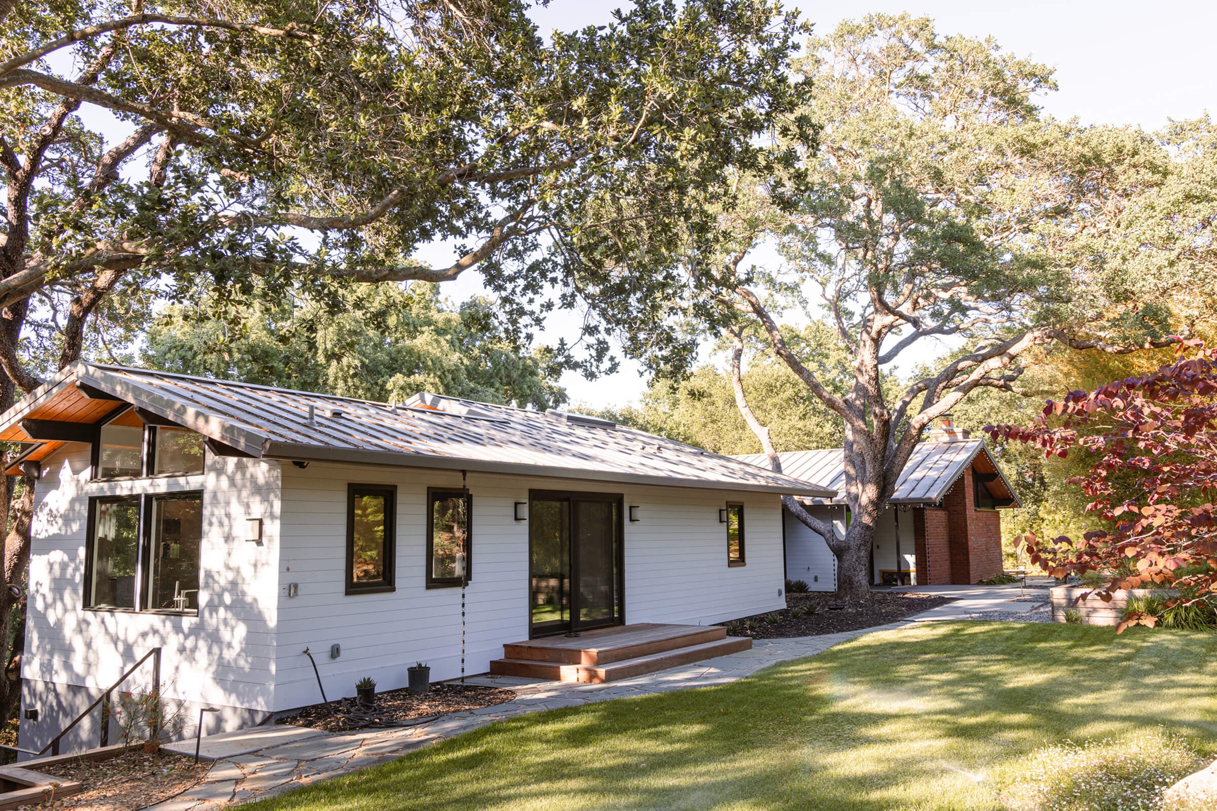 Side of the remodeled home with painted horizontal siding and a standing seam roof under oak trees